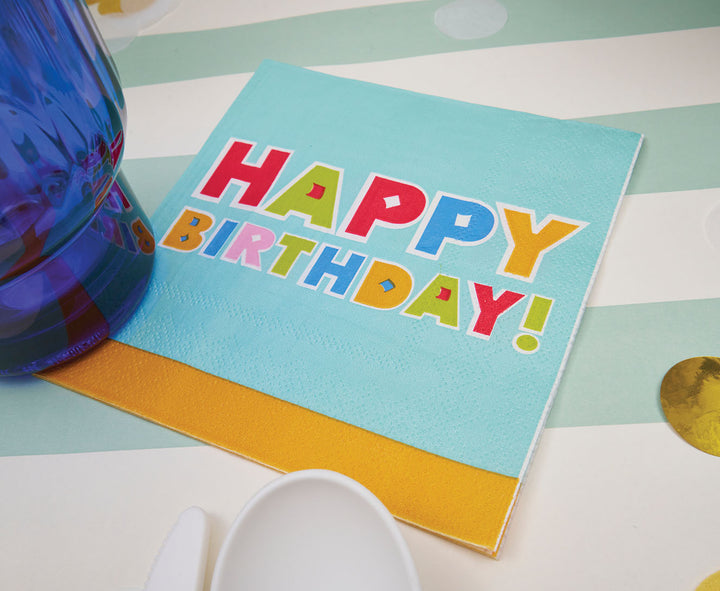 The Sophistiplate ‘Happy Birthday’ Cocktail Napkins (26pk), featuring bold, multicolored lettering, add the perfect finishing touch to your birthday table decor when placed next to a blue glass and white utensils.