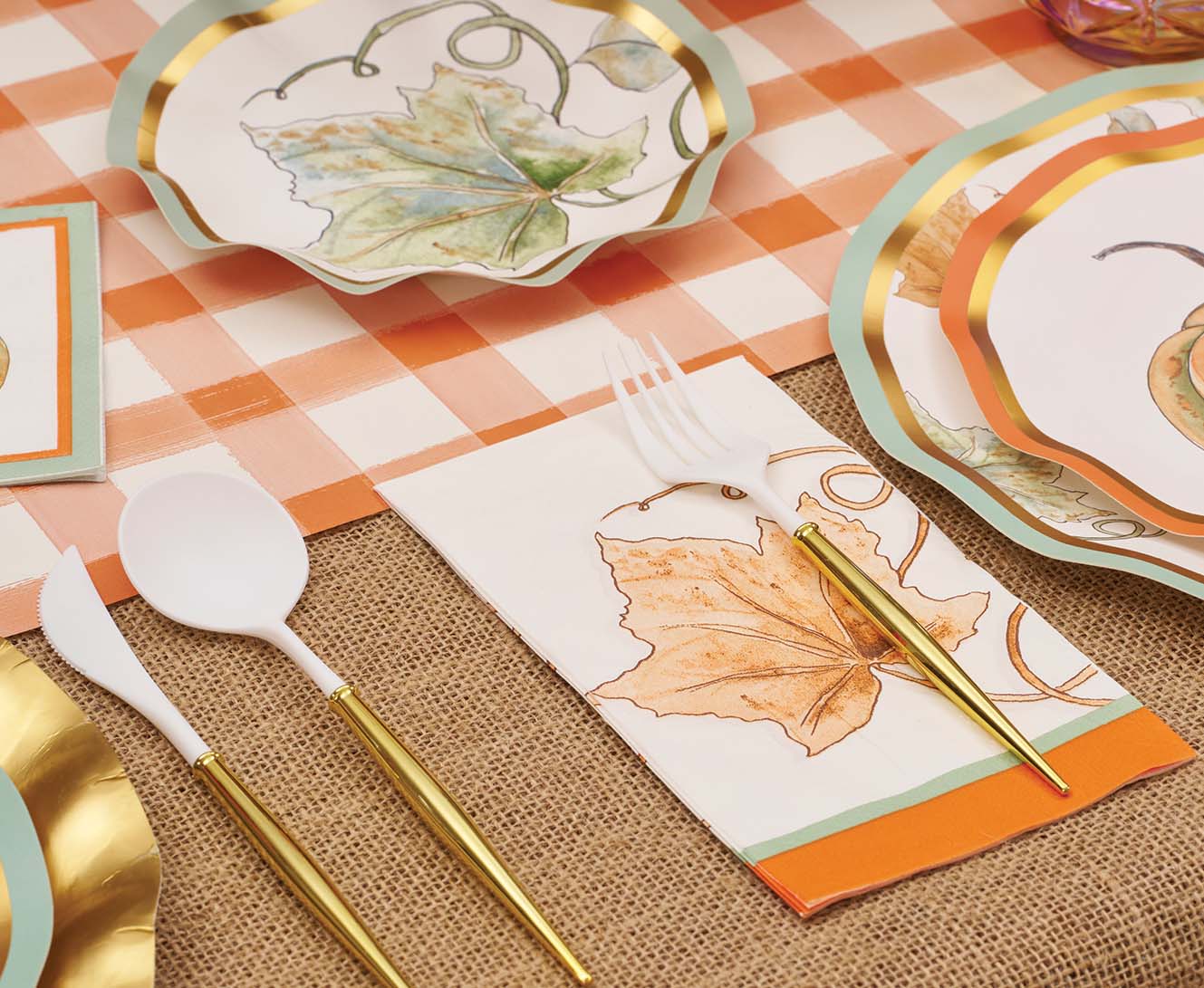 Create a fall-themed tablescape with Sophistiplate's Bright & Thankful Table Setting—designer disposable plates, napkins, and cutlery with gold accents and leaf motifs, arranged on a checkered cloth and burlap runner.