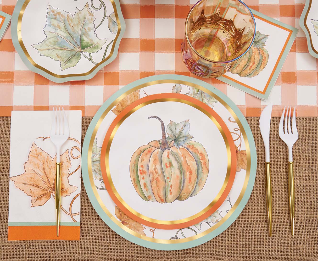 A fall-themed table setting featuring Sophistiplate’s Bright & Thankful Salad Plates (8pk), wavy paper plates with gold trim, a leaf napkin, gold utensils, a glass, and autumn leaf decorations arranged on an orange checkered tablecloth and burlap placemat.