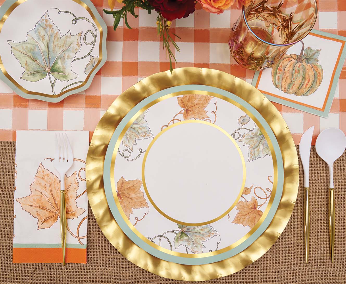 A festive fall table setting featuring Sophistiplate’s Bright & Thankful Dinner Plates (8pk), designed with metallic gold trim and pumpkin and leaf motifs, paired with gold utensils, a glass with a gold rim, a checked tablecloth, and a leaf-adorned napkin.
