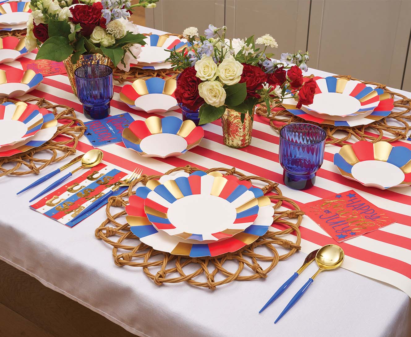 The Modern Patriotic Table Setting by Sophistiplate features red and white striped runners, designer paper plates, blue cups, gold utensils, and festive decor for a cheerful party atmosphere.