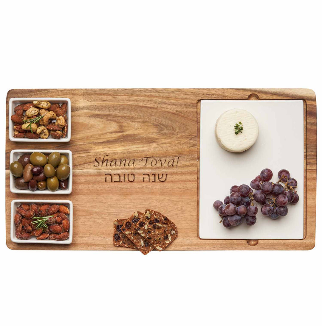 A Maple Acacia Fête Set Tray engraved with “Shana Tova” (18″ x 9.5″), beautifully arranged with ceramic snack dishes holding nuts, olives, almonds, crackers, red grapes, and a round cheese on a white serving tray.
