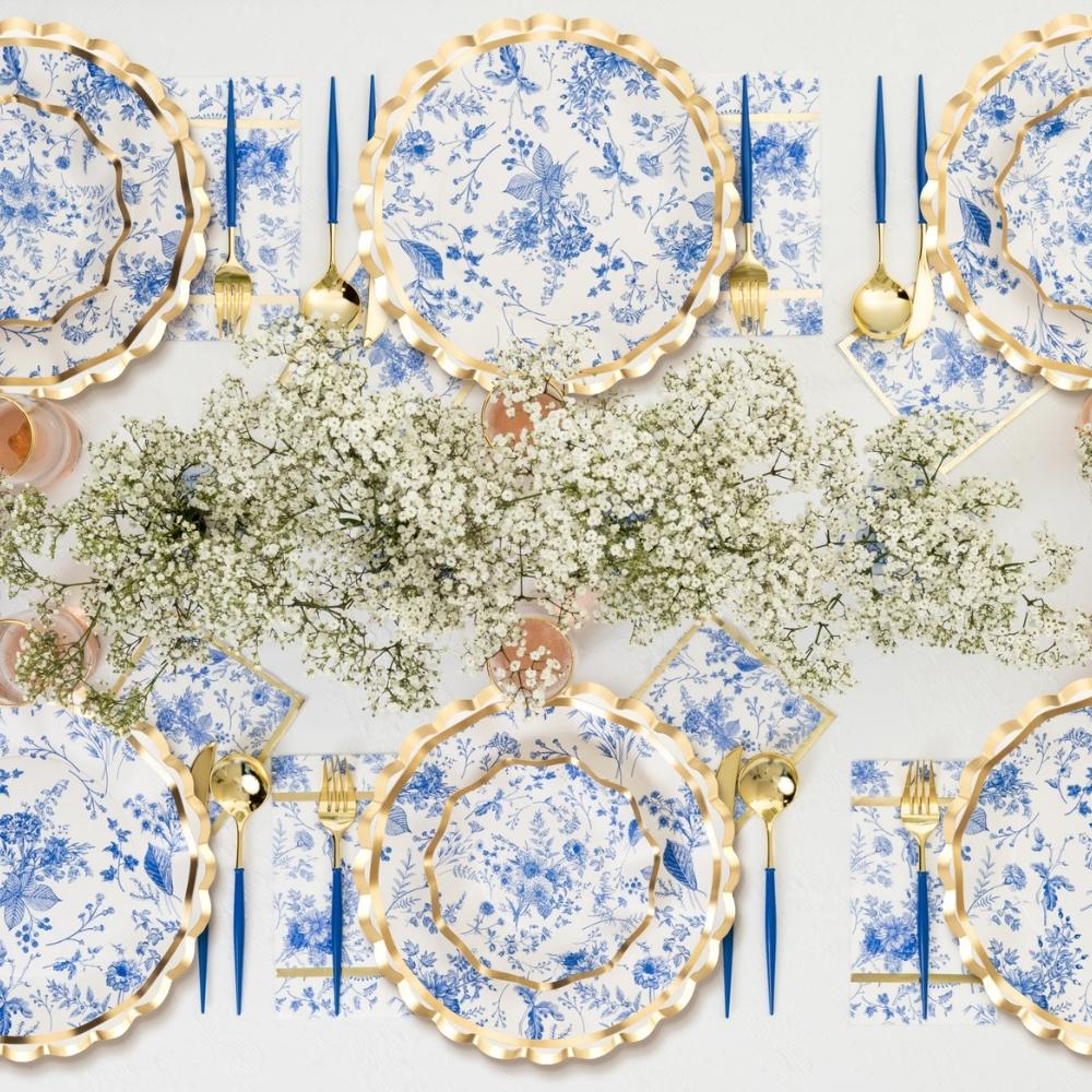 The Sophistiplate LLC Passover Tablescape for 16: Timeless features blue and white floral designer paper plates, gold-edged chargers, gold flatware, blue-handled knives, and a baby's breath centerpiece for an elegant and refined look.