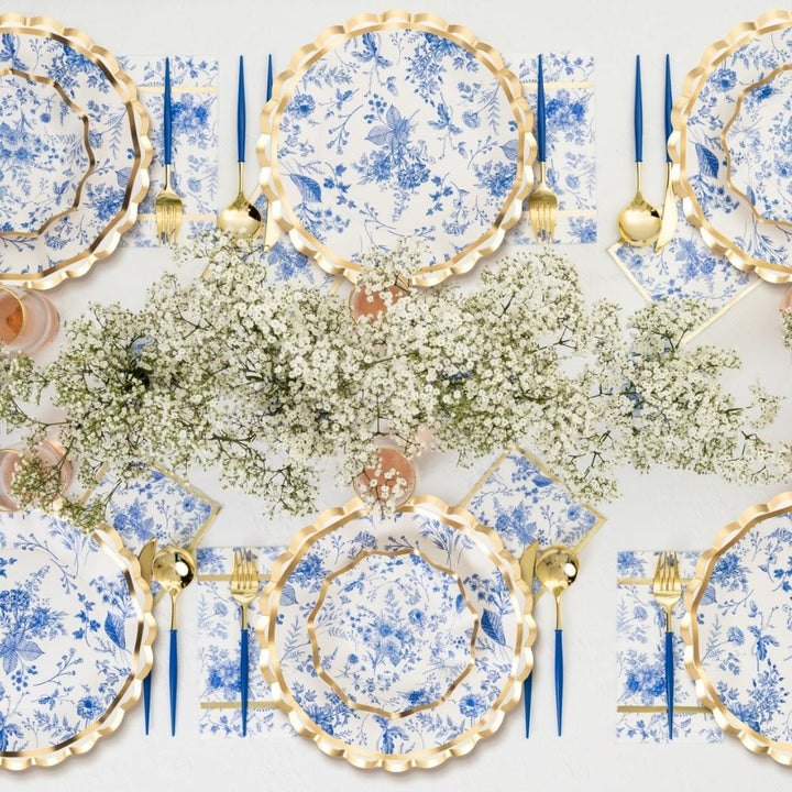 The Sophistiplate LLC Passover Tablescape for 16: Timeless features blue and white floral designer paper plates, gold-edged chargers, gold flatware, blue-handled knives, and a baby's breath centerpiece for an elegant and refined look.