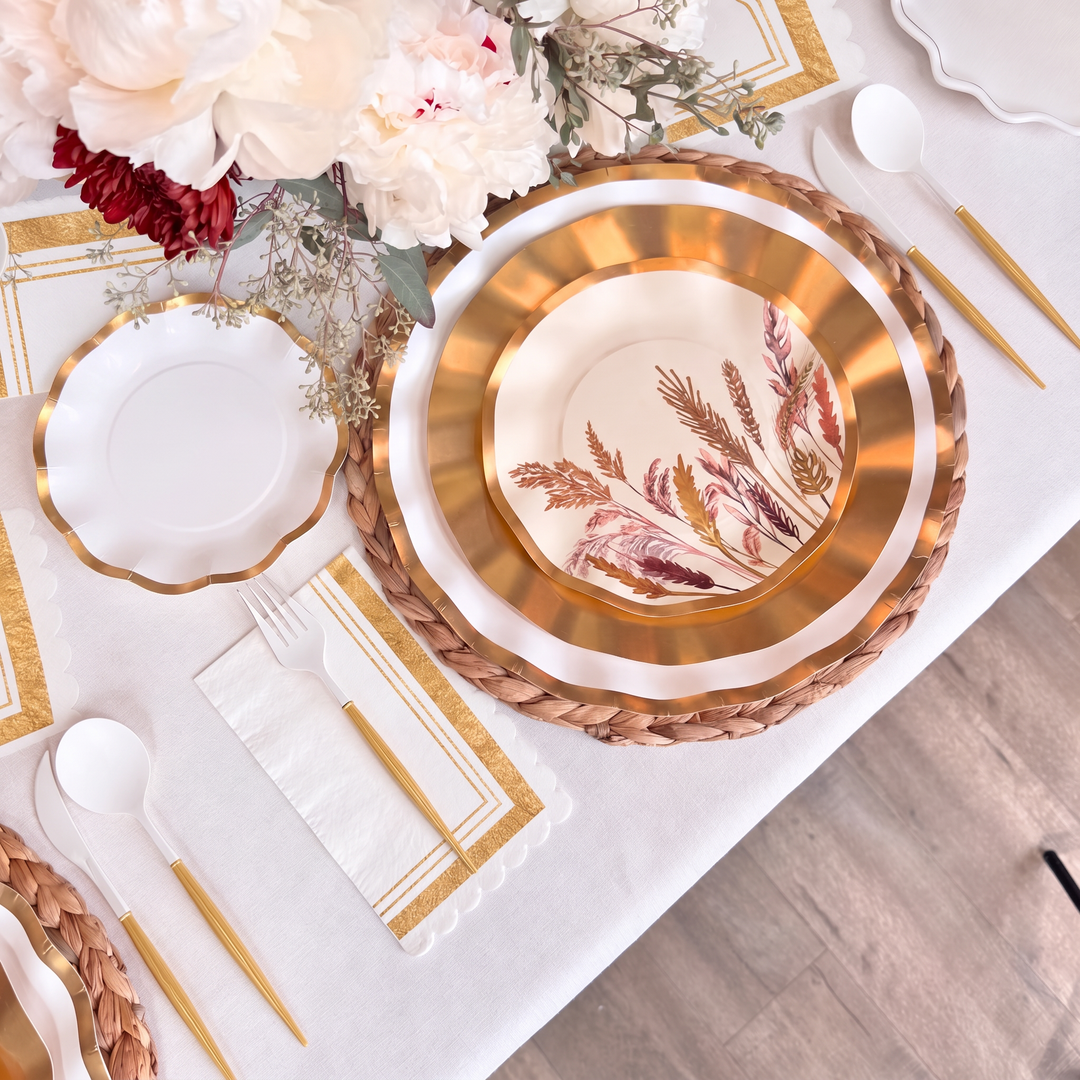 Create an elegant tablescape with the Harvest Gold Table Setting by Sophistiplate, featuring a woven placemat, gold-edged decorative paper plates, a floral-patterned bowl, gold flatware, a white napkin, and a beautiful floral centerpiece atop a crisp white tablecloth.