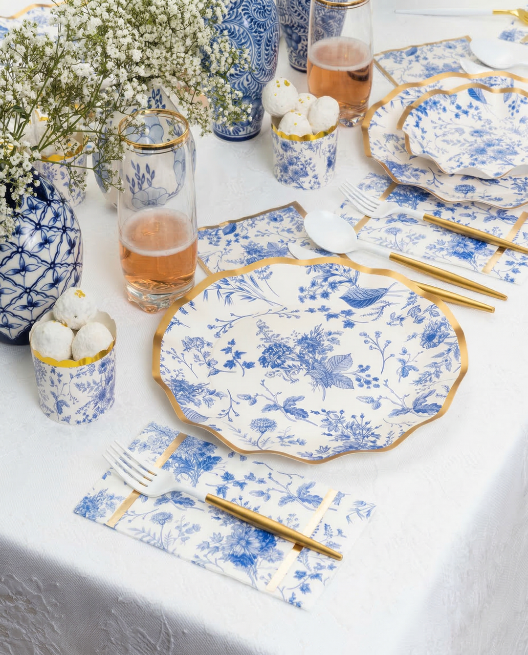 Create an elegant Passover tablescape for 16 with Sophistiplate LLC’s Timeless set, featuring blue and white floral paper plates, gold-rimmed cutlery, pink drinks in glasses, white treats in cups, and baby's breath vases on a crisp white tablecloth.