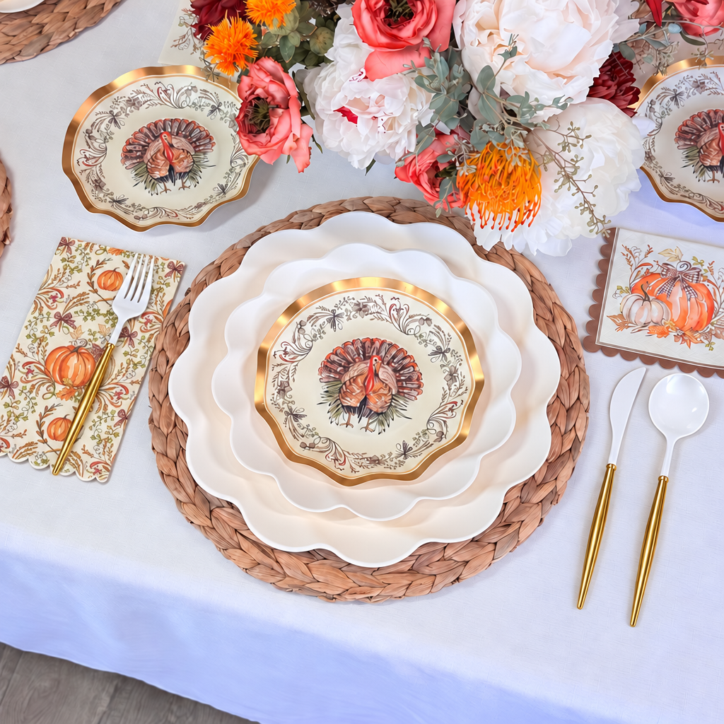 A festive table setting featuring the Ivory Harvest Table Setting by Sophistiplate, with turkey-themed plates, gold utensils, a floral centerpiece, and autumn-patterned napkins and coasters, all arranged on a woven placemat atop a white tablecloth.