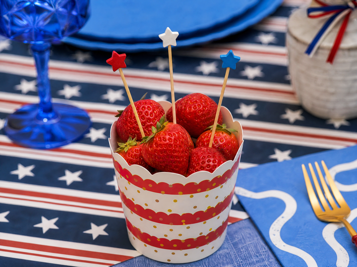 A cup of fresh strawberries with red, white, and blue star picks sits on a patriotic table featuring stars, stripes, festive napkins, and eco-friendly plates from the 4th of July Table for 16+: French Blue by Sophistiplate LLC.