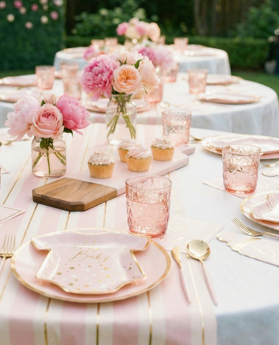 The Hey Baby Blush Table Setting by Sophistiplate features pink and gold decor, baby onesie-shaped designer plates, pink glassware, gold cutlery, cupcakes, and vases of pink flowers on a striped table runner for a perfect baby shower.