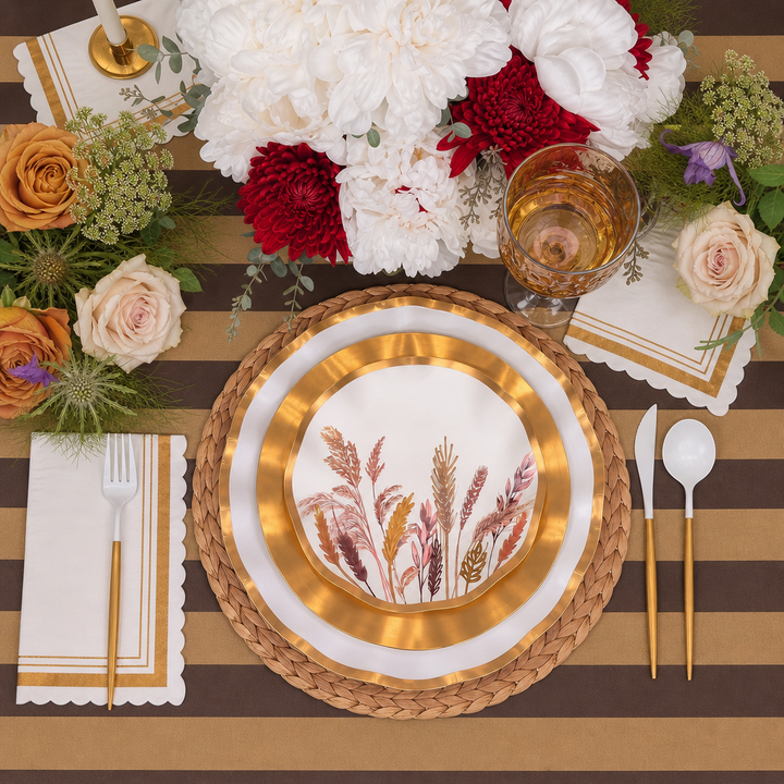 The Harvest Gold Table Setting by Sophistiplate features a gold-bordered decorative paper plate with elegant floral designs, complemented by gold cutlery, a glass of white wine, and a floral centerpiece. The tablescape in a box is completed with a tablecloth adorned in brown and beige stripes.