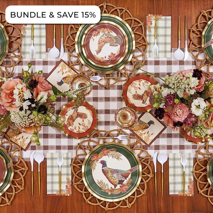 A Woodland Harvest: Thanksgiving Table for 16 set with pheasant and autumn leaf paper plates, napkins, woven chargers, gold-handled cutlery, and floral centerpieces on a brown plaid tablecloth.