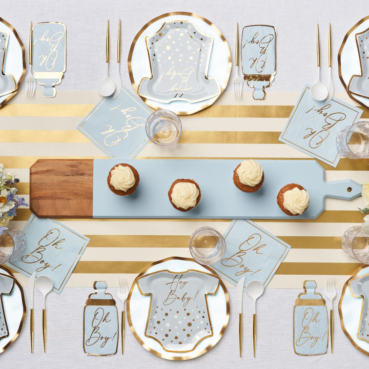 The Hey Baby Blue Table Setting by Sophistiplate LLC features a gold and white striped runner, blue and gold onesie-shaped plates, “Oh Boy!” napkins, cupcakes on a blue tray, plus clear cups and utensils—perfect for a baby shower.
