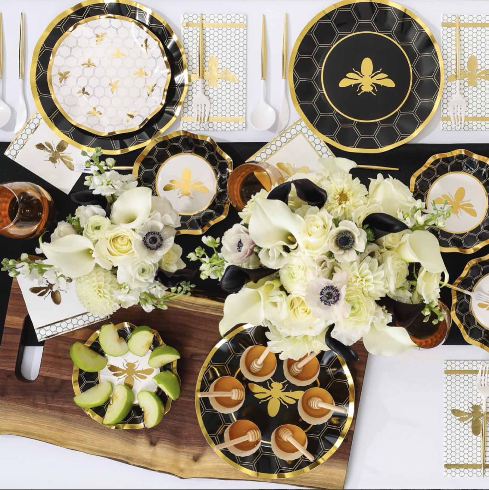 A party table set with Sophistiplate Honeybee Paper Cocktail Napkins, black, white, and gold bee-themed plates and cups, gold cutlery, honeycomb patterns, floral centerpieces, and apple slices on a wooden tray.