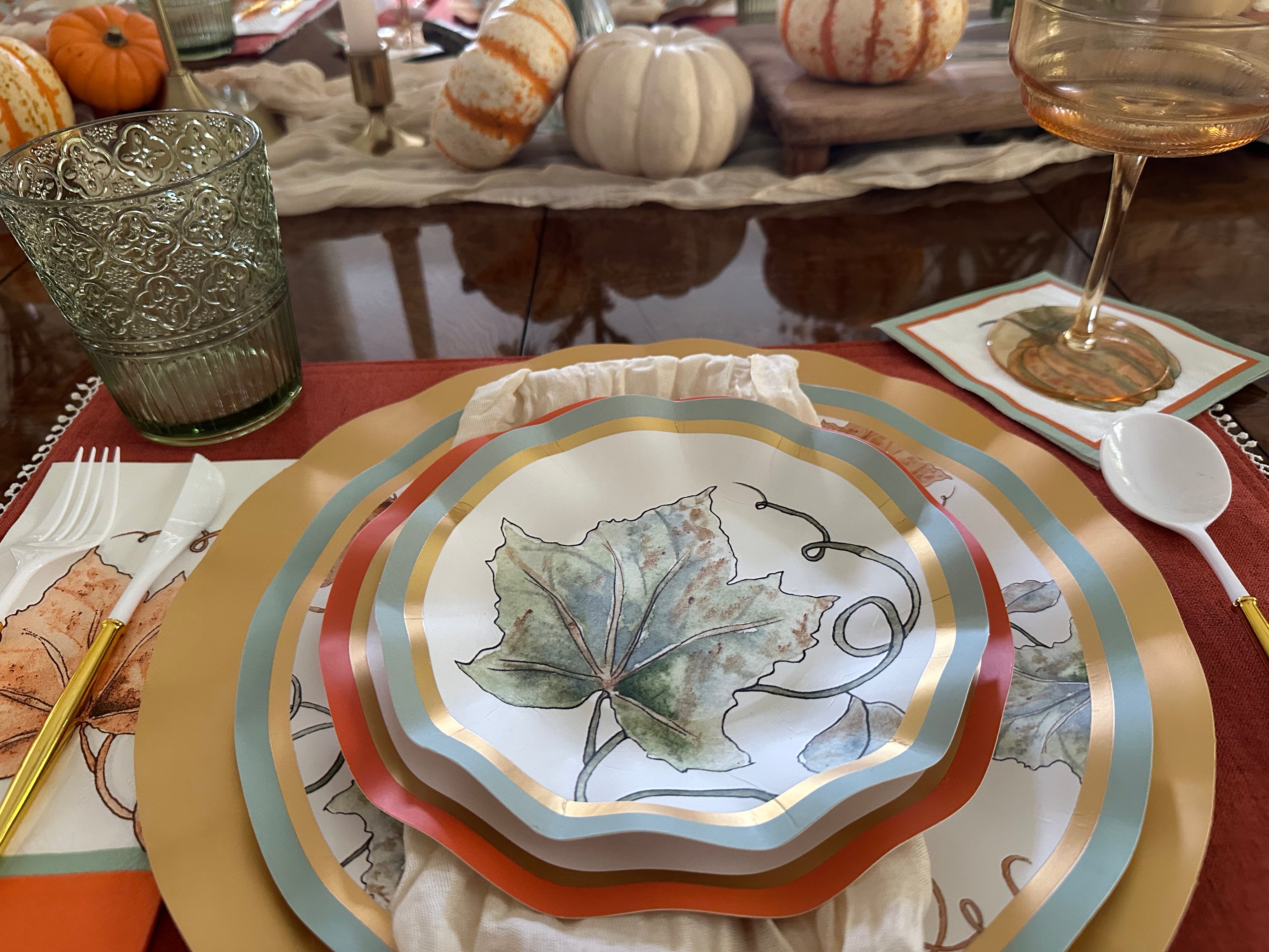 The Bright & Thankful Table Setting by Sophistiplate showcases layered pumpkin harvest plates with leaf designs, gold accents, green glassware, utensils, and candles, plus decorative pumpkins arranged along the center for a festive autumn look.