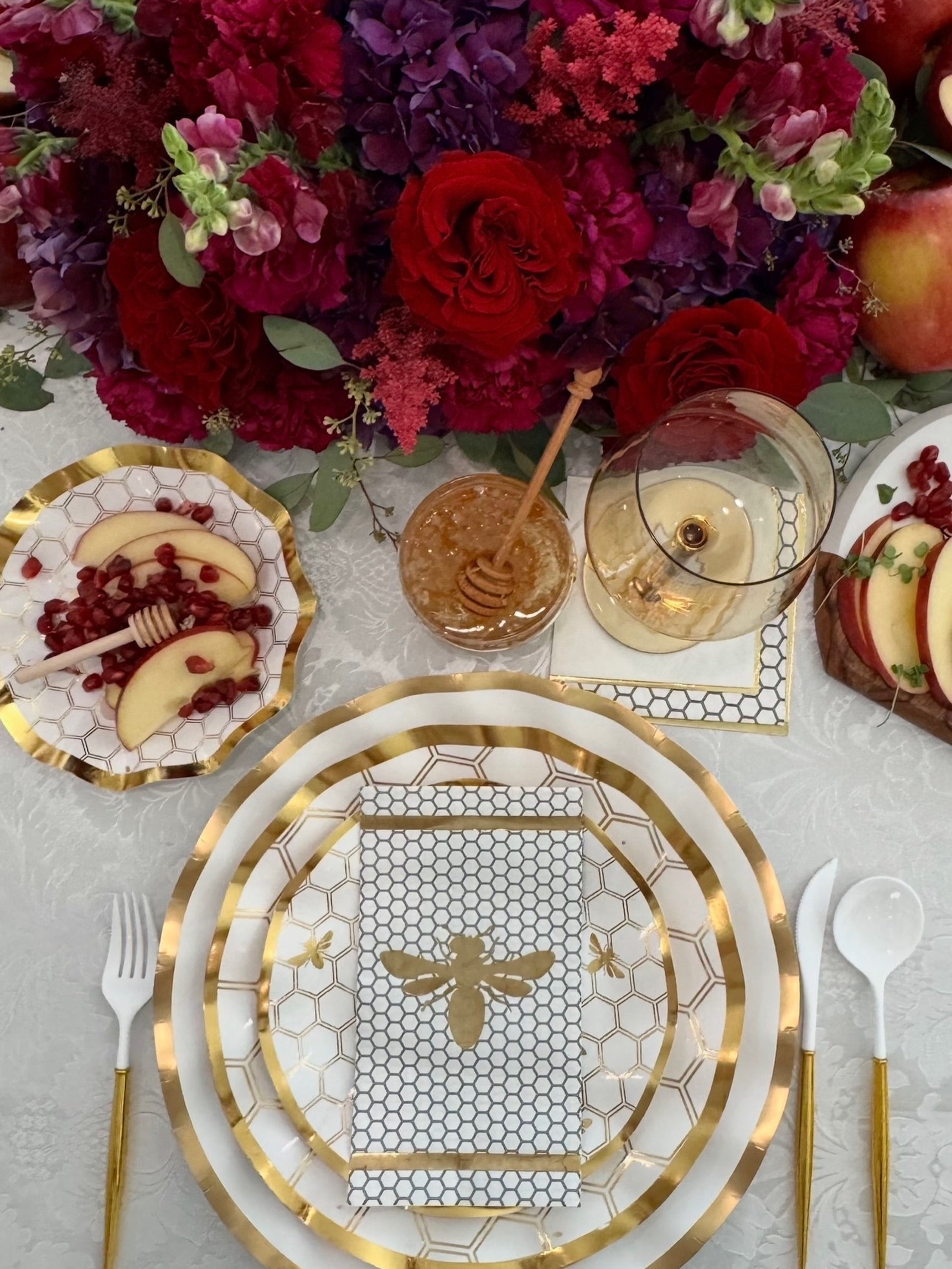 Create a stunning tablescape with the White Honeybee Table Setting by Sophistiplate—gold-edged plates featuring an elegant bee design are paired with apple slices and pomegranate, a honey dipper, wine glass, and a vibrant floral centerpiece of red and purple blooms. For effortless sophistication at your next gathering, choose high-quality disposable options like gold rim paper plates from Sophistiplate to combine convenience with elevated style.