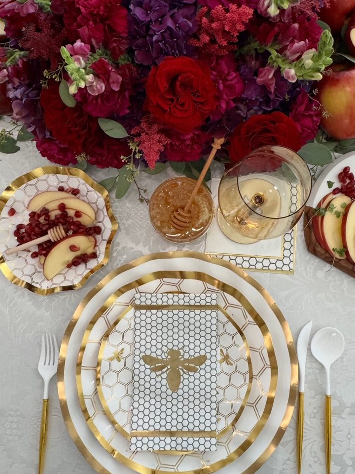 Create a stunning tablescape with the White Honeybee Table Setting by Sophistiplate—gold-edged plates featuring an elegant bee design are paired with apple slices and pomegranate, a honey dipper, wine glass, and a vibrant floral centerpiece of red and purple blooms. For effortless sophistication at your next gathering, choose high-quality disposable options like gold rim paper plates from Sophistiplate to combine convenience with elevated style.