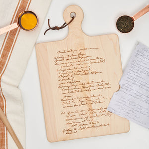 The Maple Engraved Recipe Maple Board (16 x 10) with a personalized recipe rests on a white surface next to a striped kitchen towel, two copper measuring cups with oil and herbs, and a handwritten note.