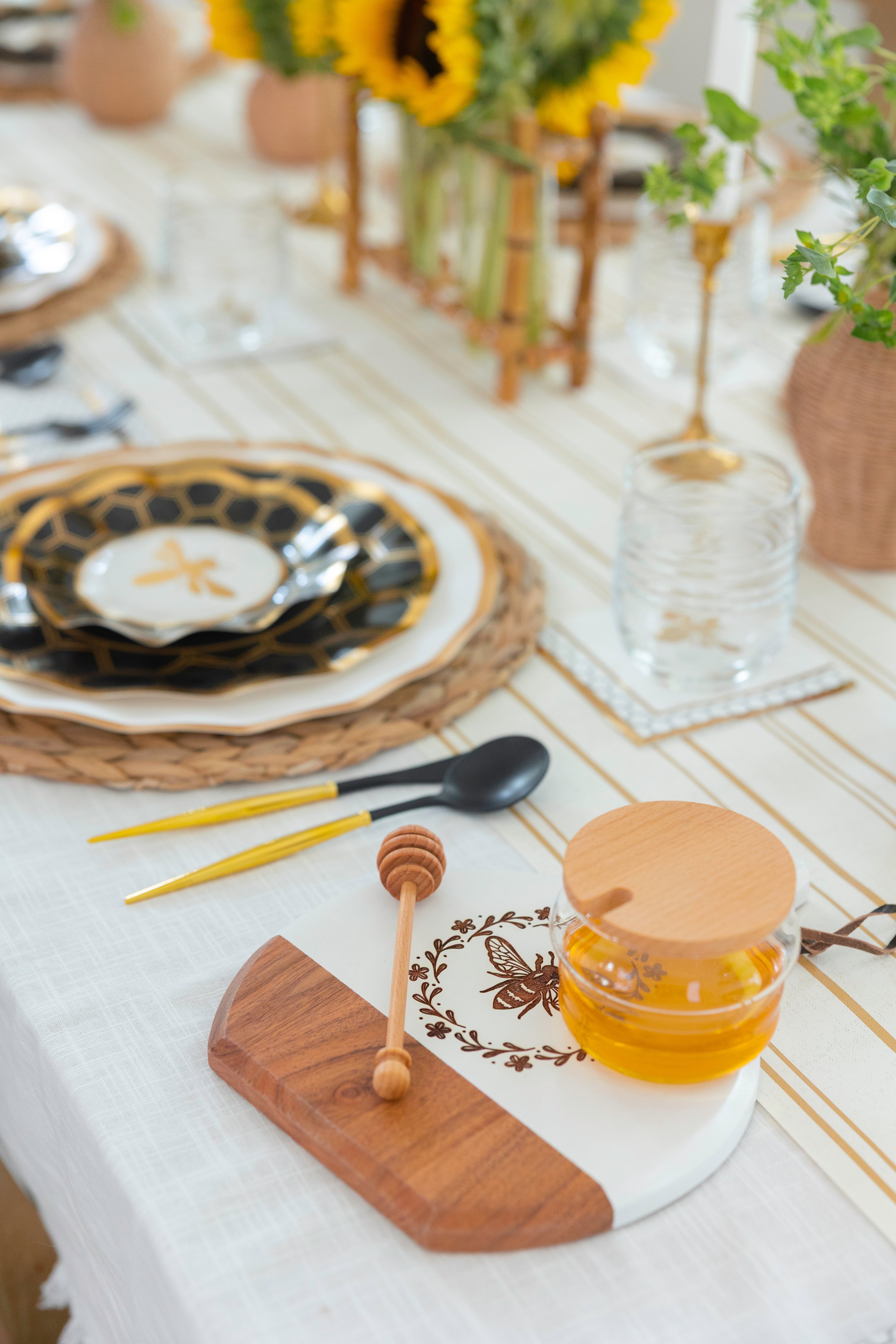The Honeybee Table Setting by Sophistiplate LLC features elegant heavy duty paper plates, gold and black utensils, a bee-themed decorative plate, honey jar with dipper, plus sunflowers and greenery for a bright, charming gathering.