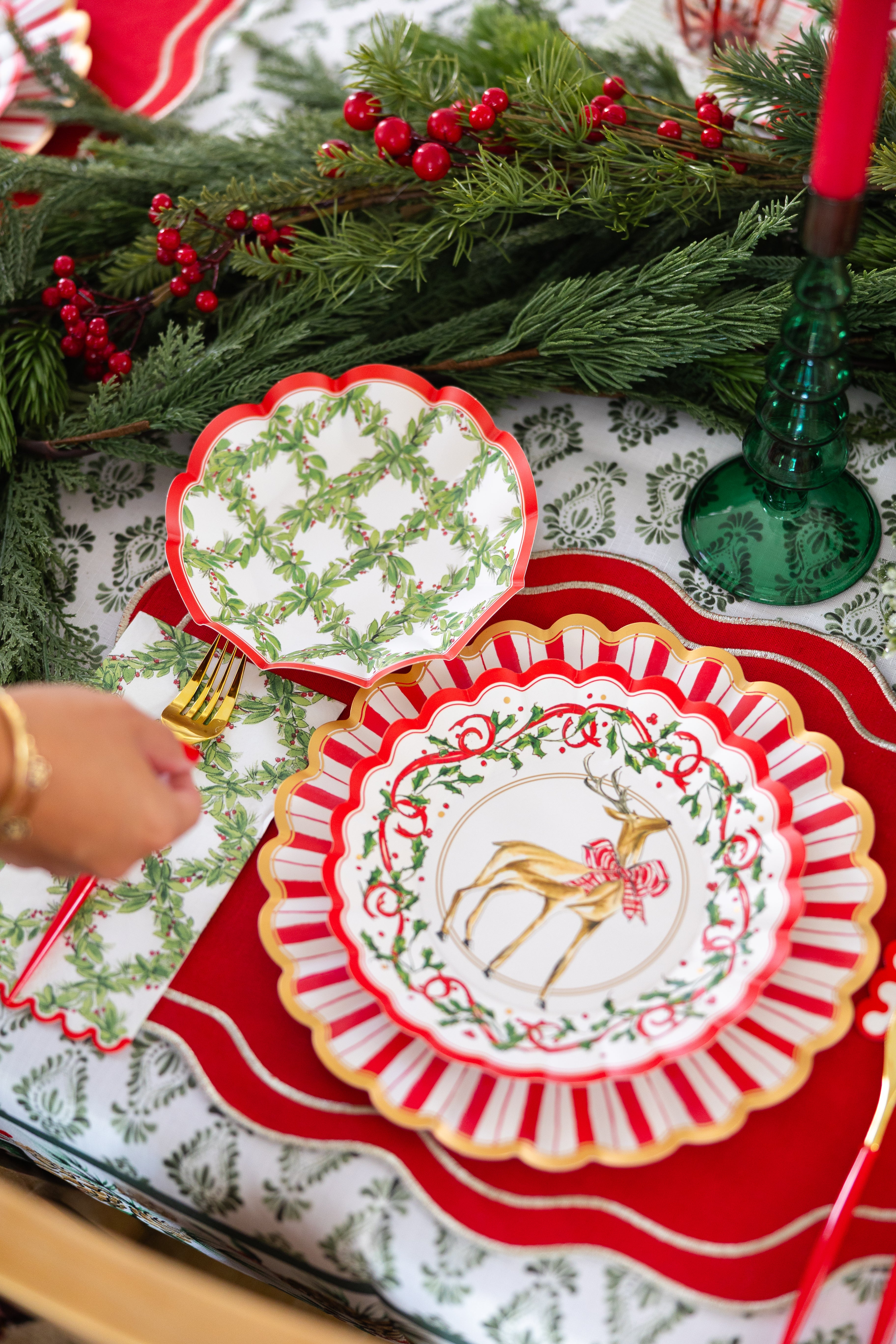 A festive holiday table set with a red placemat, gold utensils, a reindeer plate with red bow, Sophistiplate Holly Berry Guest Towels, a green and white patterned plate, and Christmas greenery with red berry garland centerpiece.