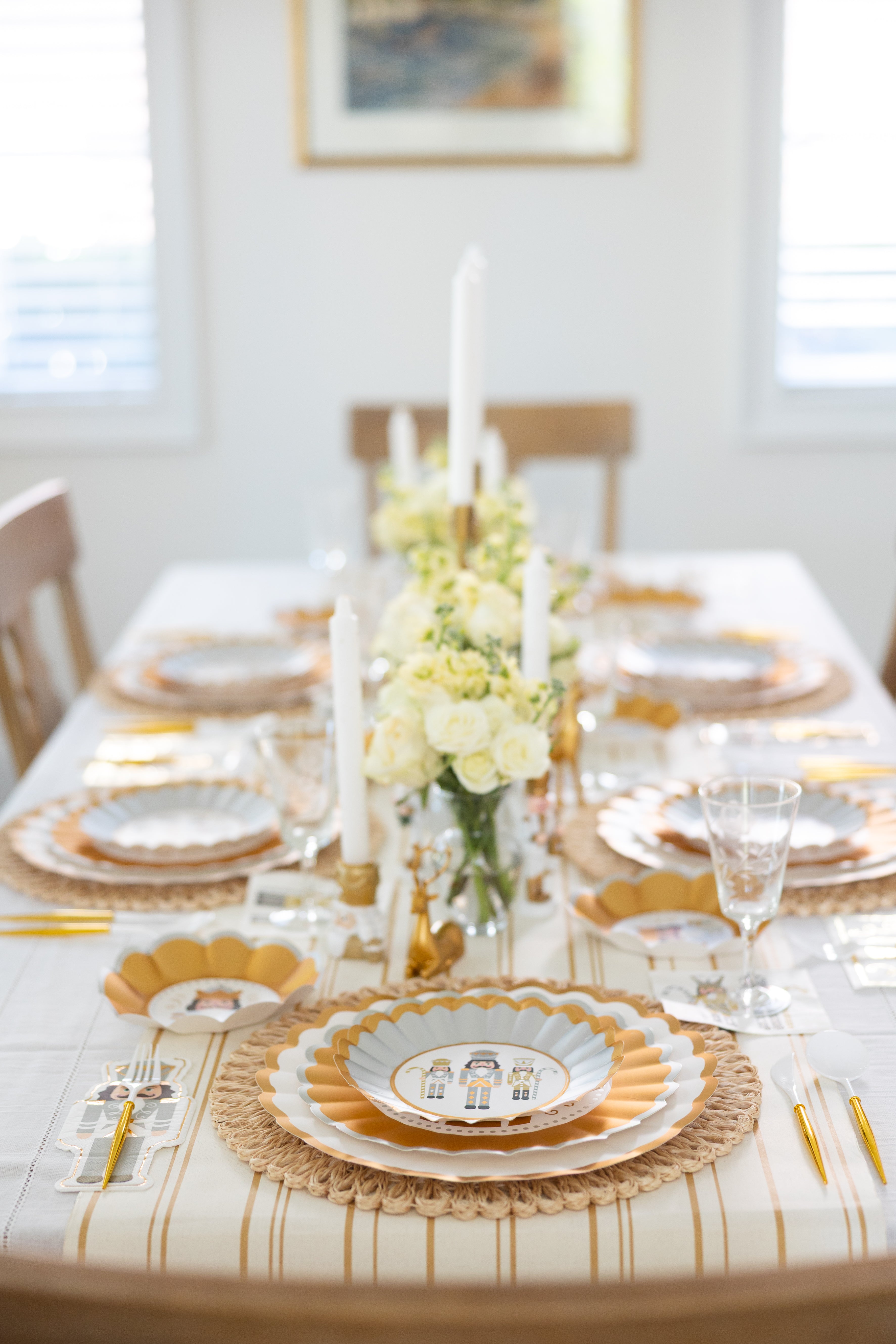 The Gilded Nutcracker Table Setting by Sophistiplate features gilded nutcracker plates, scalloped placemats, gold cutlery, clear glasses, white candles, and a centerpiece of white roses and greenery in a bright room.