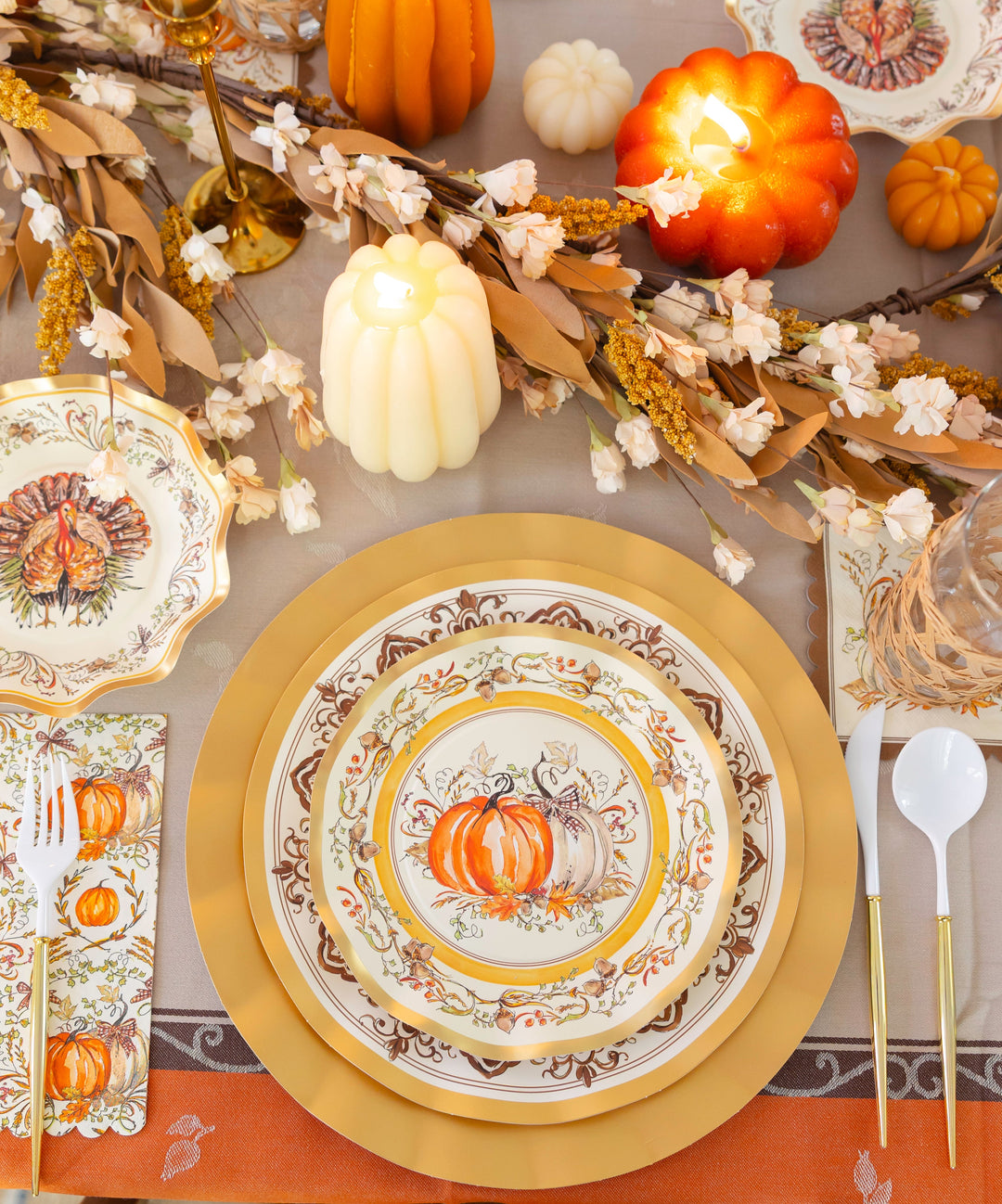 Create a warm and inviting Thanksgiving tablescape with the Turkey & Gourds Table Setting by Sophistiplate, featuring pumpkin-themed designer paper plates, gold chargers, faux pumpkins, candles, a floral garland, and vintage turkey napkins. White and gold flatware is arranged beside each place setting atop an orange tablecloth for a festive autumn look.