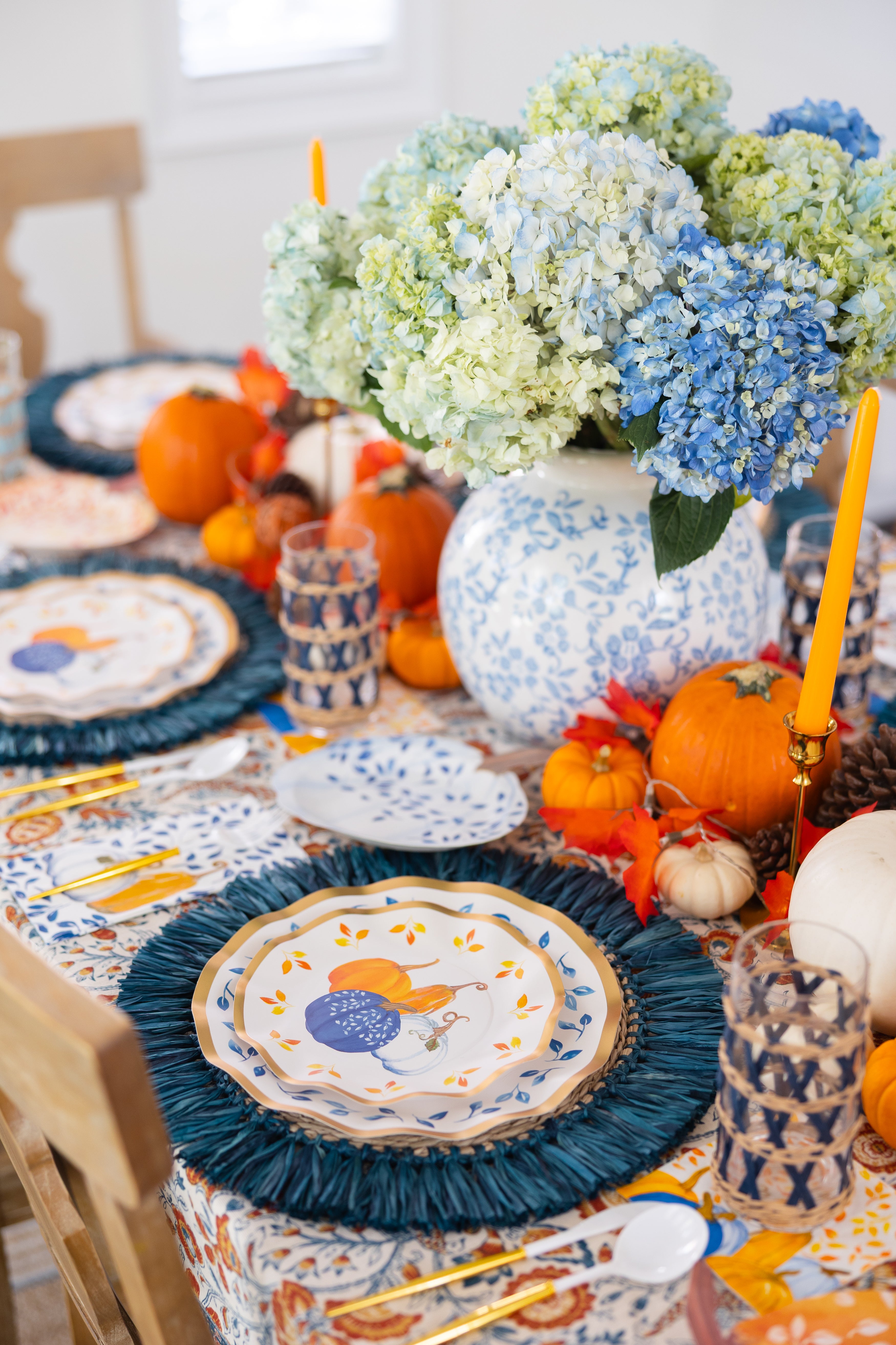 The Harvest Blues Table Setting by Sophistiplate includes blue and white plates, orange and blue glassware, small pumpkins, orange candles, hydrangeas in a vase, all arranged on a patterned tablecloth for a festive look.