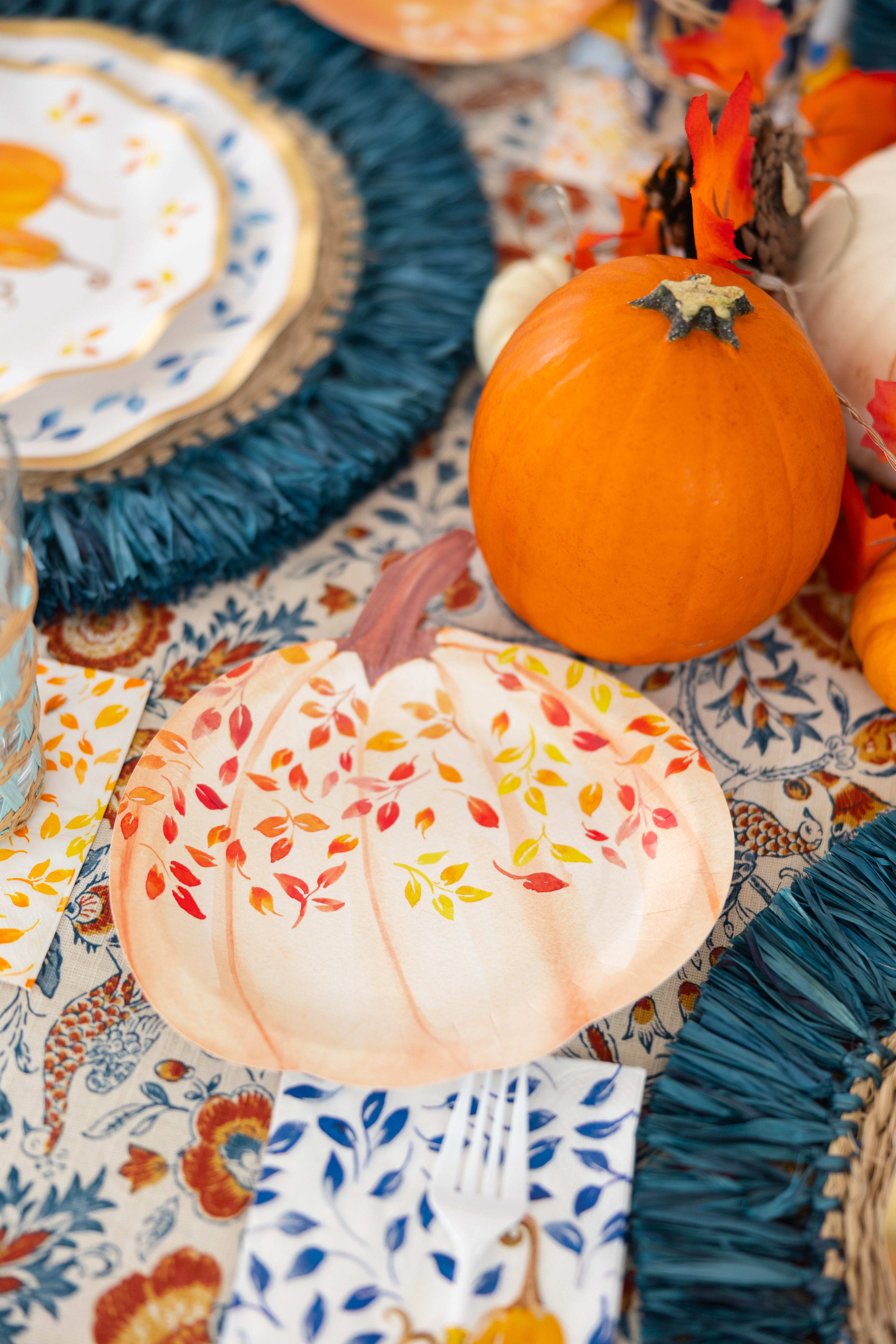 The Sophistiplate Harvest Blues Table Setting includes a small pumpkin, a leafy pumpkin-shaped designer plate, blue fringe placemats, and floral napkins with blue and yellow leaves on a patterned tablecloth.