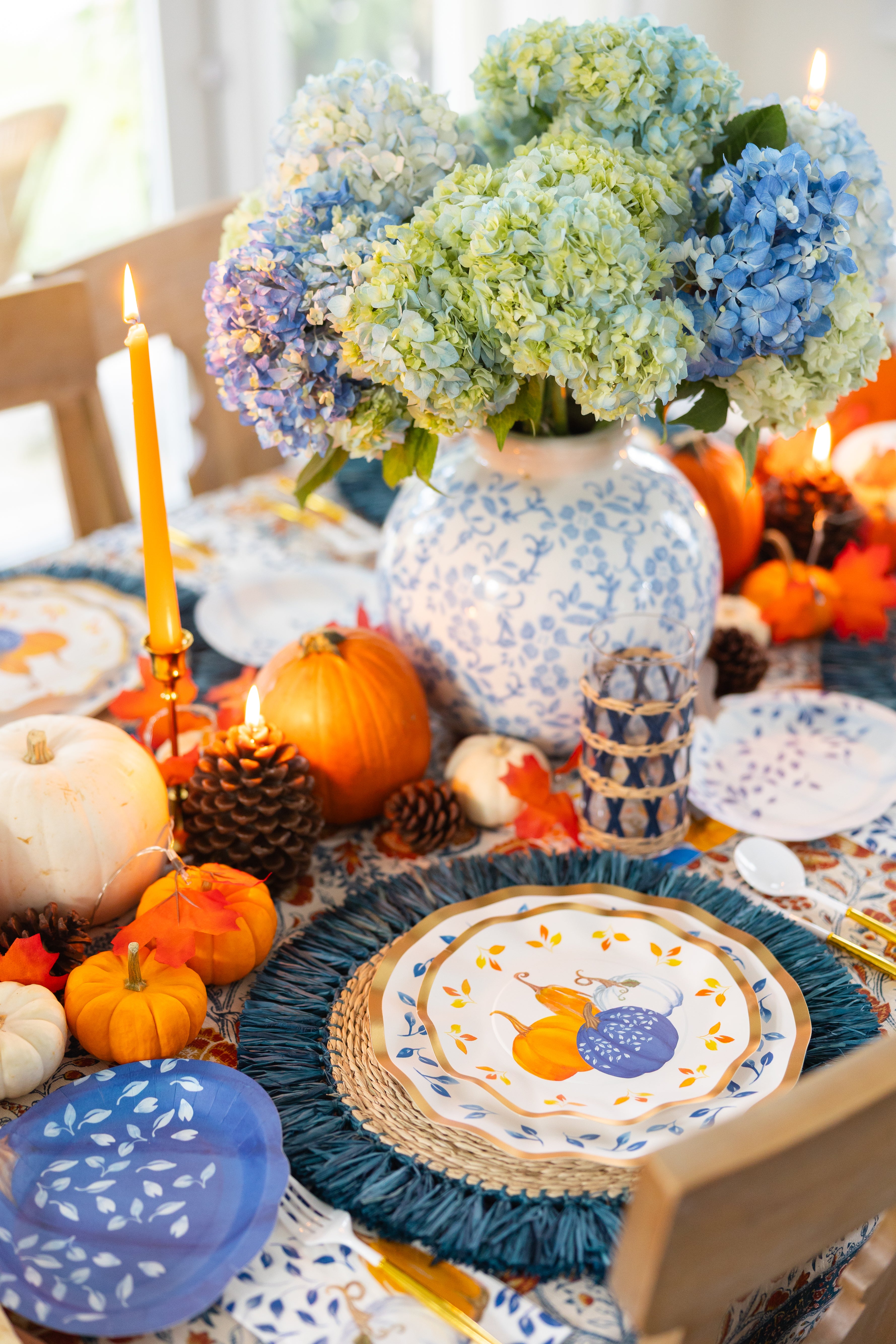 The Harvest Blues Table Setting by Sophistiplate features designer blue and orange plates, a blue hydrangea centerpiece, pumpkins, pinecones, candles, and fall leaves for an easy autumn tablescape-in-a-box.