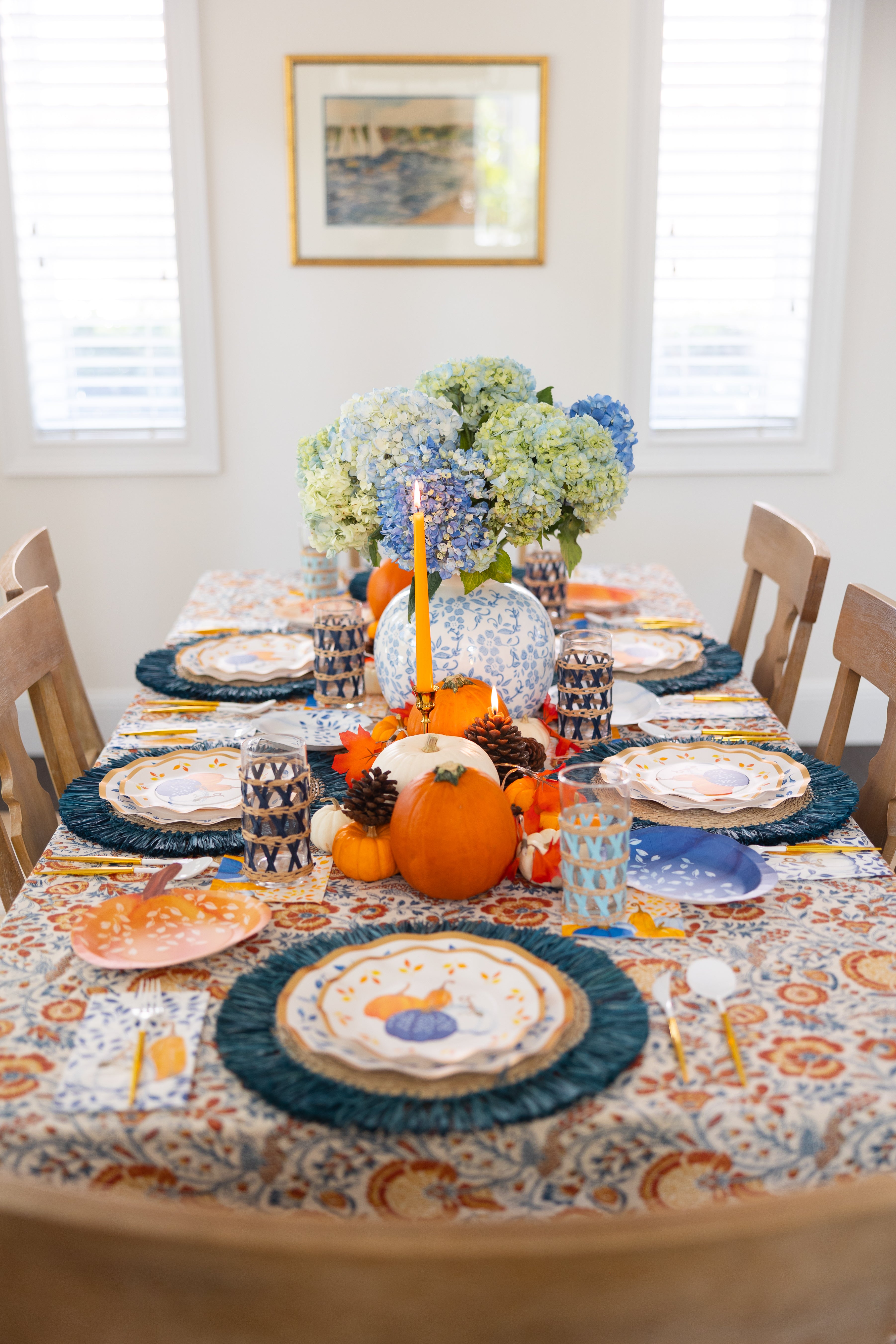 The Harvest Blues Table Setting by Sophistiplate showcases designer paper plates, blue placemats, orange candles, pumpkins, pinecones, blue hydrangeas, and decorative cups on a vibrant tablecloth in a sunlit room.