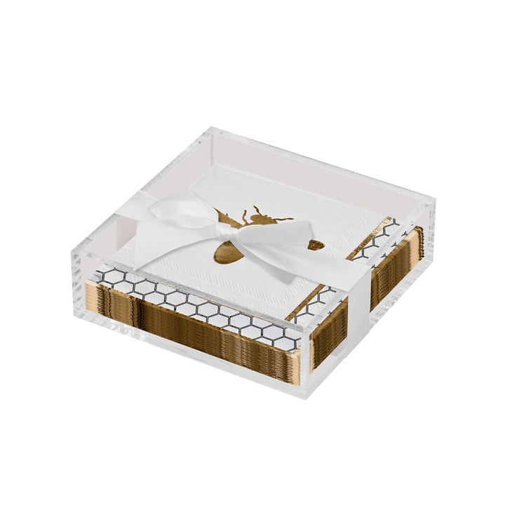 The Sophistiplate Honeybee Cocktail Napkins with Acrylic Tray features white cocktail napkins adorned with a gold bee motif, elegantly arranged in a clear acrylic tray with a honeycomb pattern and tied with a white ribbon—an ideal addition to any modern tabletop.