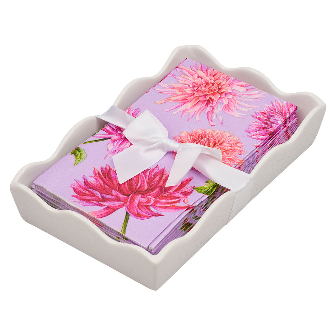 The Sophistiplate Floral Guest Towel with Napkin Tray features purple napkins with pink flowers, neatly stacked in a white wavy-edged ceramic holder and tied with a white ribbon bow.