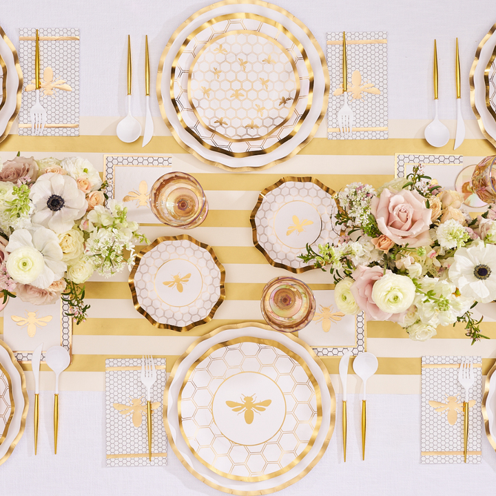 A White Honeybee Table Setting by Sophistiplate, featuring gold and white party plates adorned with elegant bee and honeycomb designs, gold cutlery, floral centerpieces, and striped yellow and white table runners, creates a stunning atmosphere—showing how even sophisticated paper plates can add charm and sophistication to your celebration.