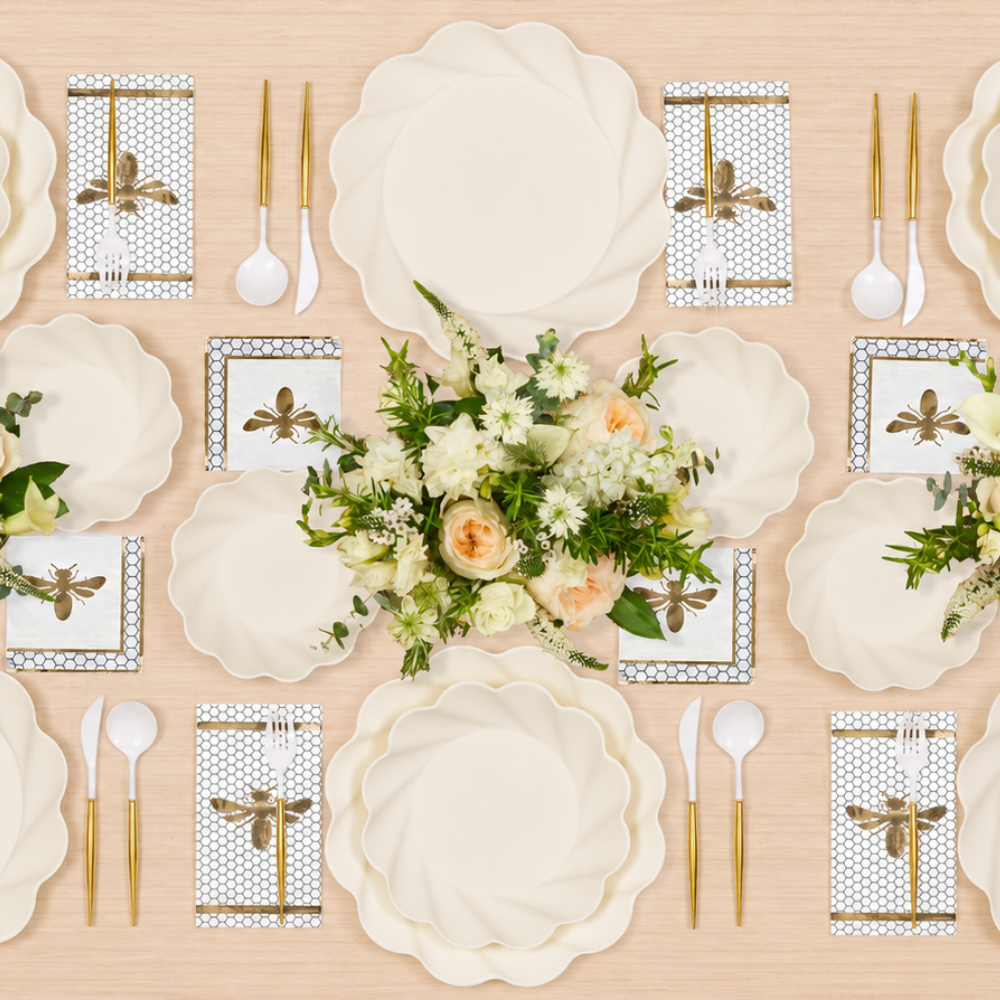 An overhead view showcases the Sophistiplate Eco Cream Honeybee Table Setting, featuring elegant paper plates, gold cutlery, white napkins adorned with gold bees, and a floral centerpiece of white and peach flowers with greenery—creating a stunning “tablescape in a box.”.