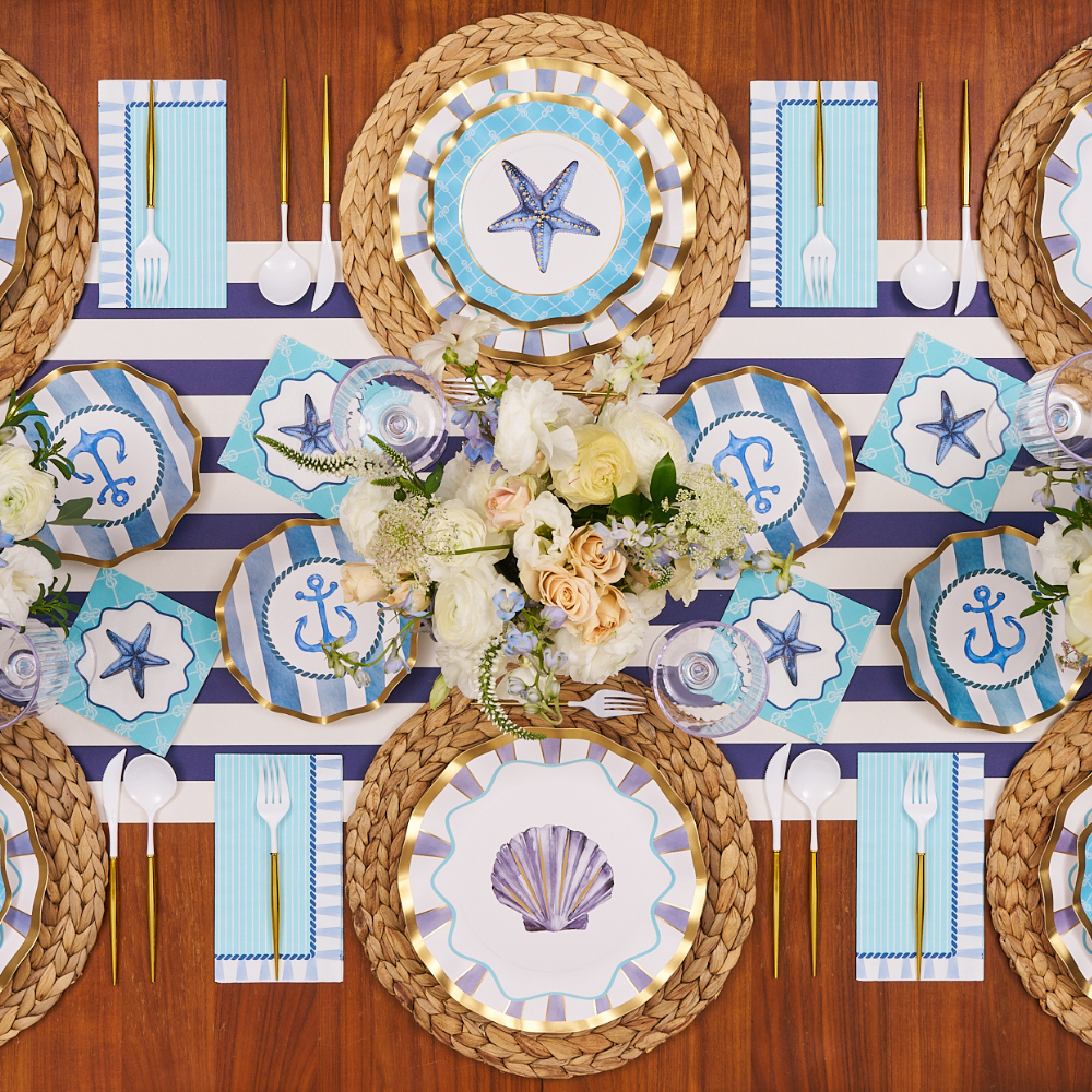 Create a nautical-themed tablescape with the Sophistiplate Tidal Wave Table Setting, featuring seashell, starfish, and anchor motifs, coordinating blue-and-white accents, gold utensils, and woven placemats.