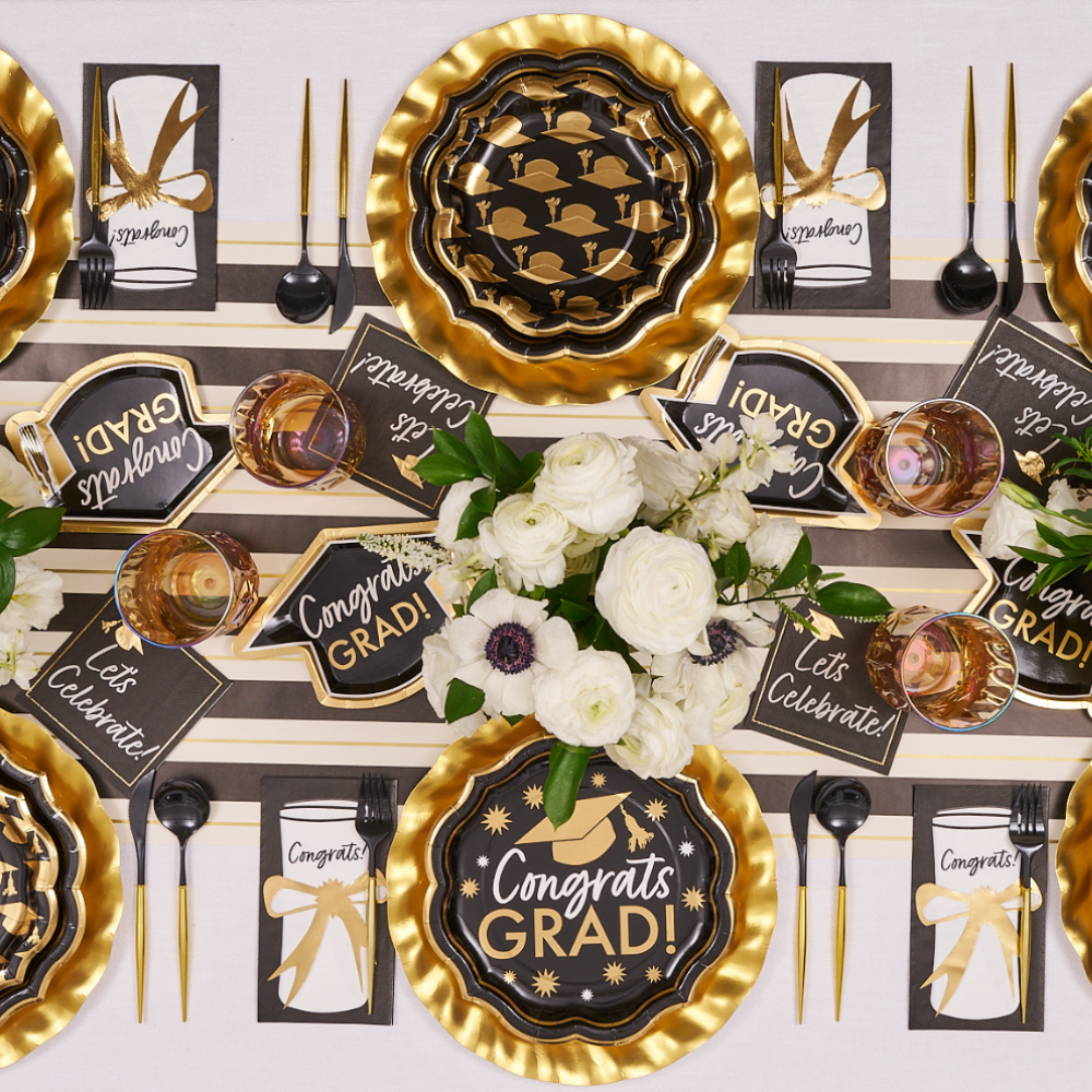 The Sophistiplate Classic Grad Table Setting features black and gold paper plates, “Congrats Grad!” signs, gold utensils, bow-tied napkins, and white floral centerpieces—perfect for stylish graduation party decor.