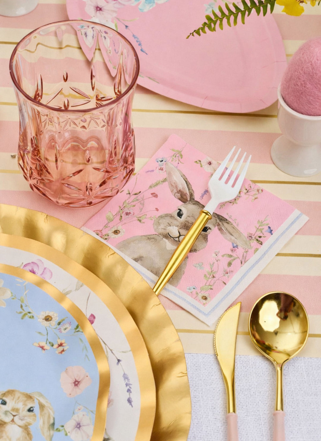 The Ultimate Easter Tablescape for 16: Charming Easter by Sophistiplate LLC features pastel designer paper plates, a pink glass, gold utensils, a bunny napkin, pink egg holder, and heart-shaped plate for festive spring flair.