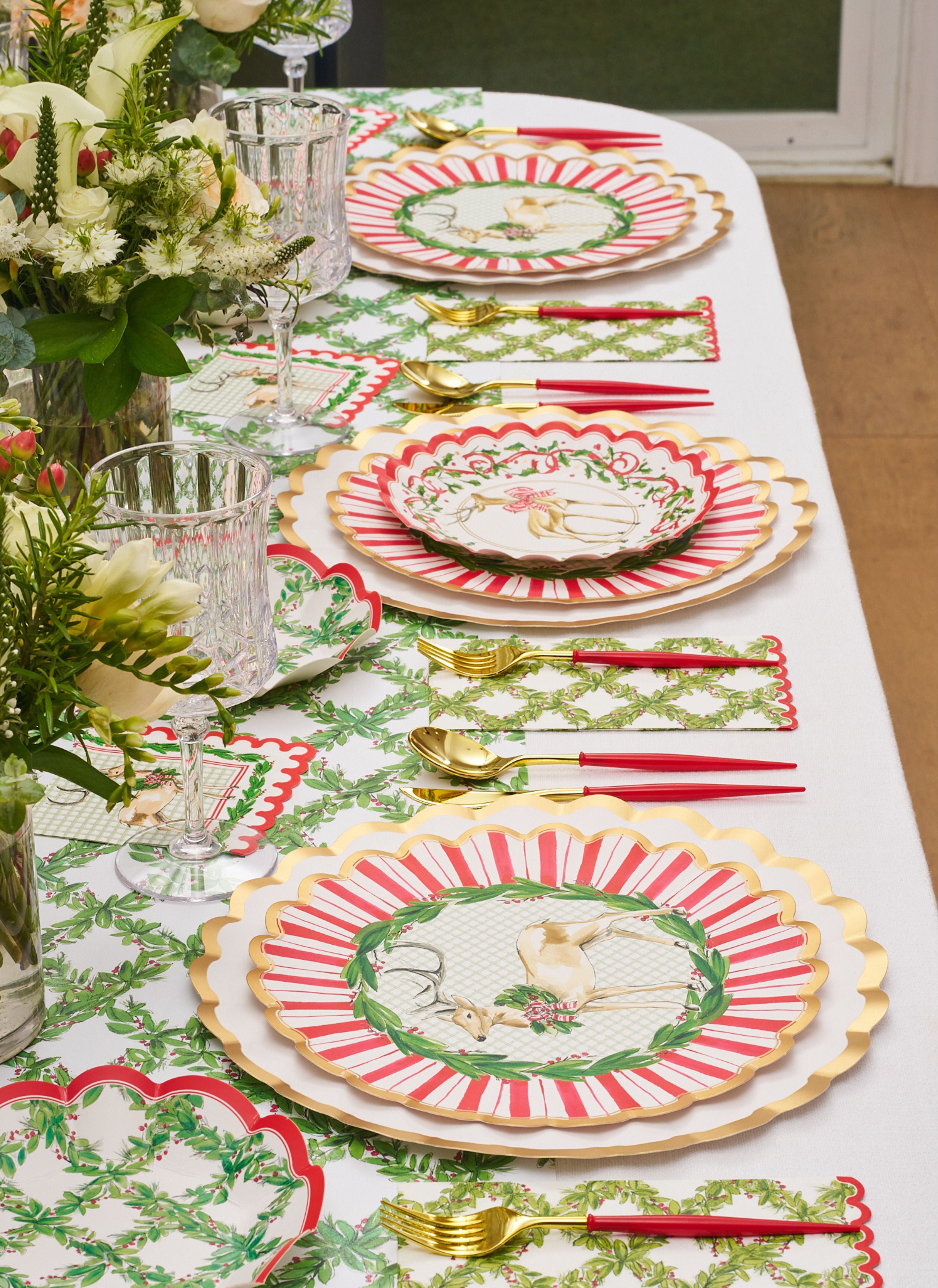 A festive table is set with gold-rimmed plates, red and white stripes, floral and bird designs, gold cutlery with red handles, crystal glasses, and the Rosanne Beck Holly Berry Trellis Table Runner (20x12