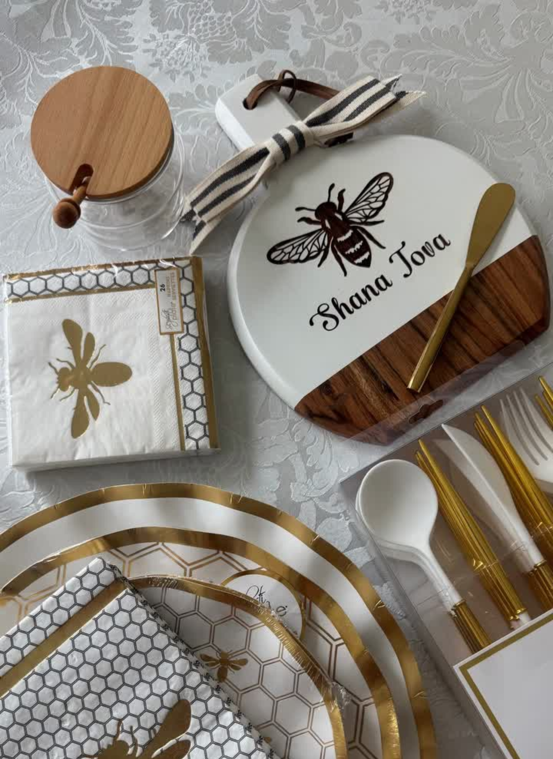 A Rosh Hashanah table set with bee-themed plates, napkins, utensils, a honey jar, and the Maple Bevel Board with Spreader Knife—engraved with “Shana Tova” and a bee graphic (9.75 x 8”)—all arranged on a white patterned tablecloth.