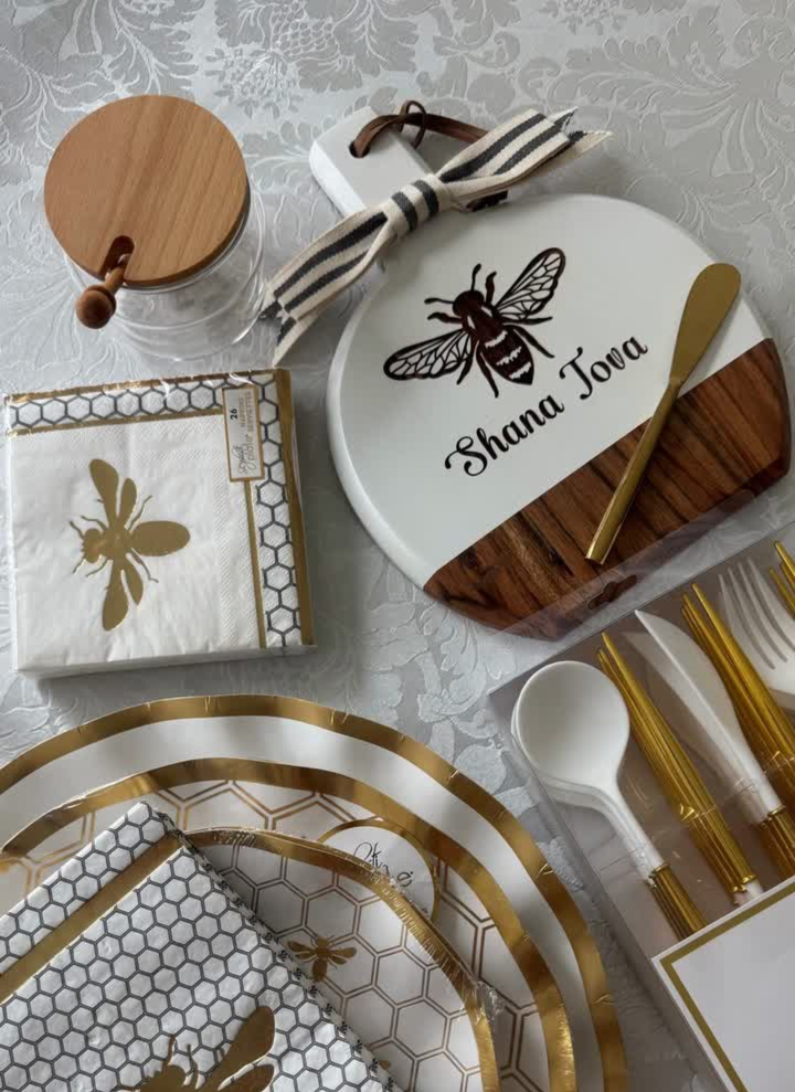 A Rosh Hashanah table set with bee-themed plates, napkins, utensils, a honey jar, and the Maple Bevel Board with Spreader Knife—engraved with “Shana Tova” and a bee graphic (9.75 x 8”)—all arranged on a white patterned tablecloth.