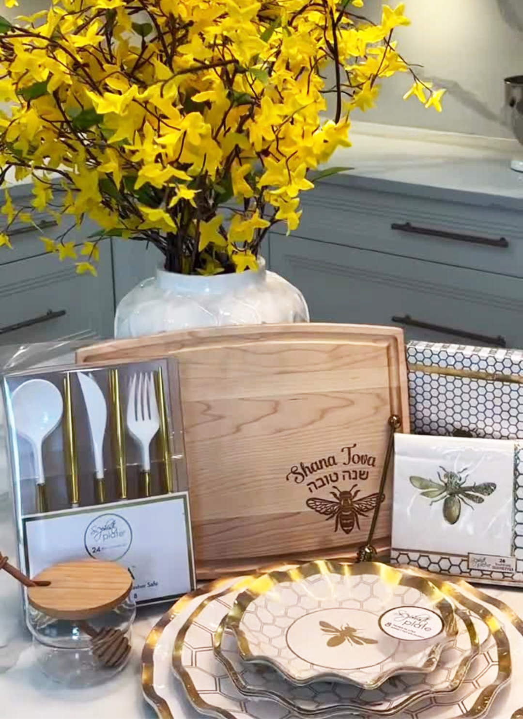 A Rosh Hashanah table setting featuring bee-themed plates, napkins, utensils, and yellow flowers in a vase, all arranged on a kitchen counter alongside a Maple 12″ x 9″ Arched Maple Board engraved with “Shana Tova.”.
