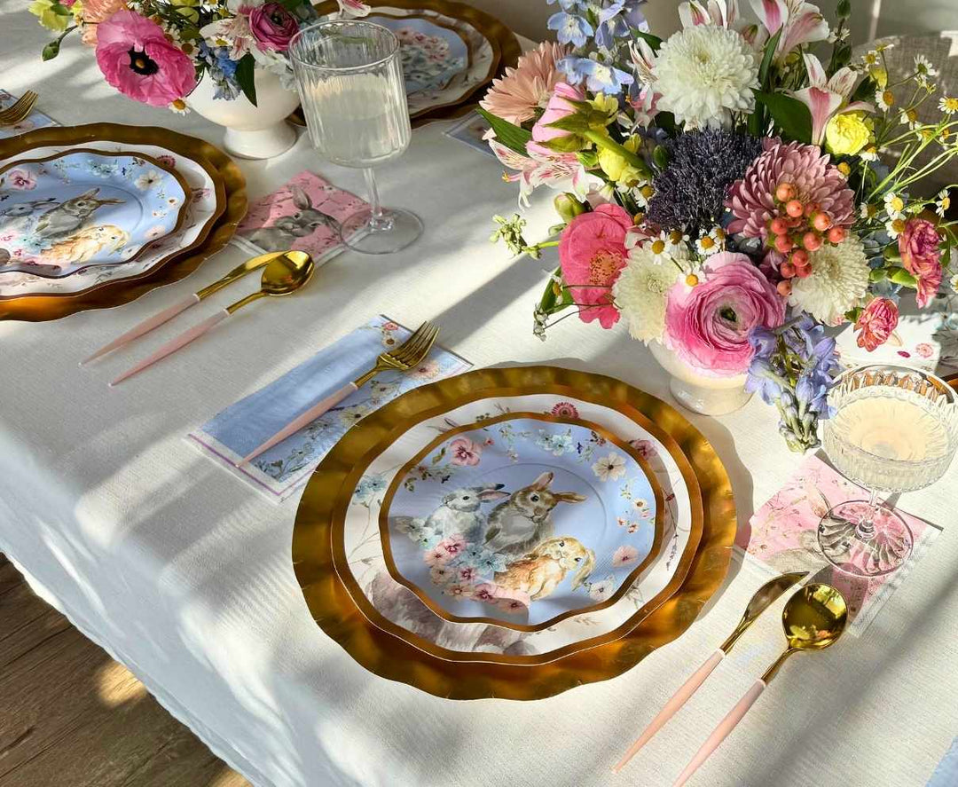 Create a spring-themed table with Sophistiplate LLC's Ultimate Easter Tablescape for 16: Charming Easter, featuring designer plates with rabbits and lambs, pastel pink cutlery, floral napkins, wine glasses, and flower décor on a white tablecloth.
