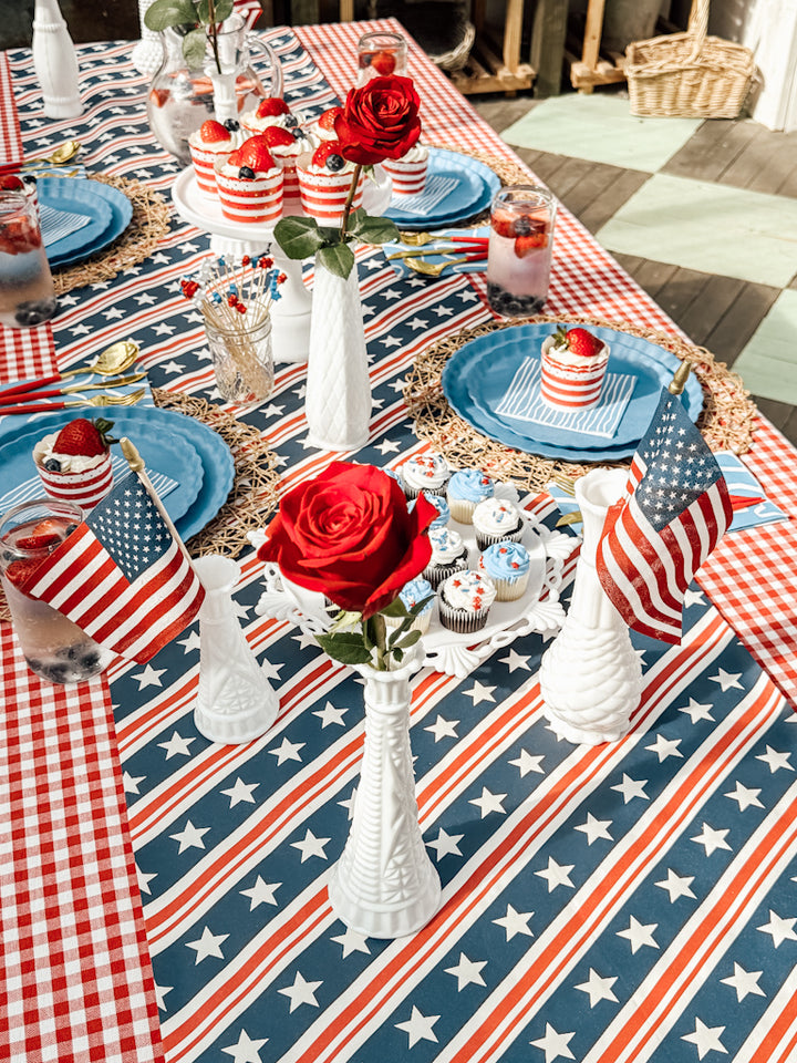 Celebrate with a festive Patriotic French Blue table setting by Sophistiplate LLC, featuring blue plates, star-spangled tablecloth, flag accents, red roses in white vases, eco-friendly disposables, and patriotic desserts for a lively atmosphere.