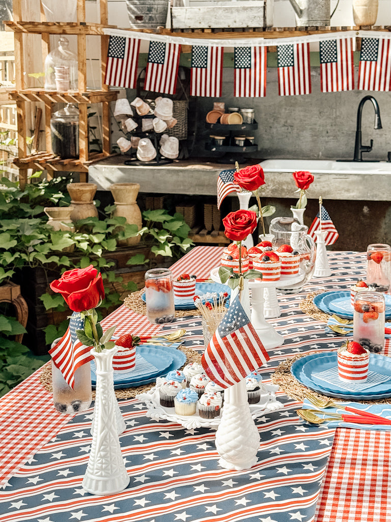 A patriotic table setting features Sophistiplate LLC's Patriotic French Blue plates, American flag-themed tablecloths, red roses in white vases, U.S. flag bunting, small flags, and desserts with strawberry drinks.