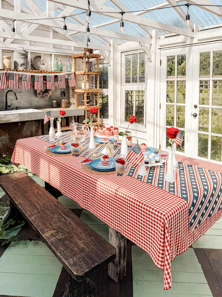 A festive table is set in a sunroom, featuring Sophistiplate LLC’s Patriotic French Blue collection with American flags, a red gingham tablecloth, and eco-friendly plates. Patriotic bunting on the wall enhances the celebration.