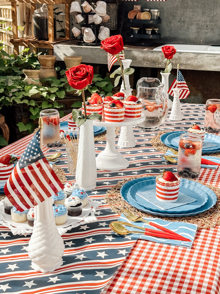 Create a festive outdoor table with Sophistiplate LLC’s Patriotic French Blue plates, American flags, red roses in white vases, layered desserts, cupcakes, fruit water, all set on a star and checkered tablecloth.