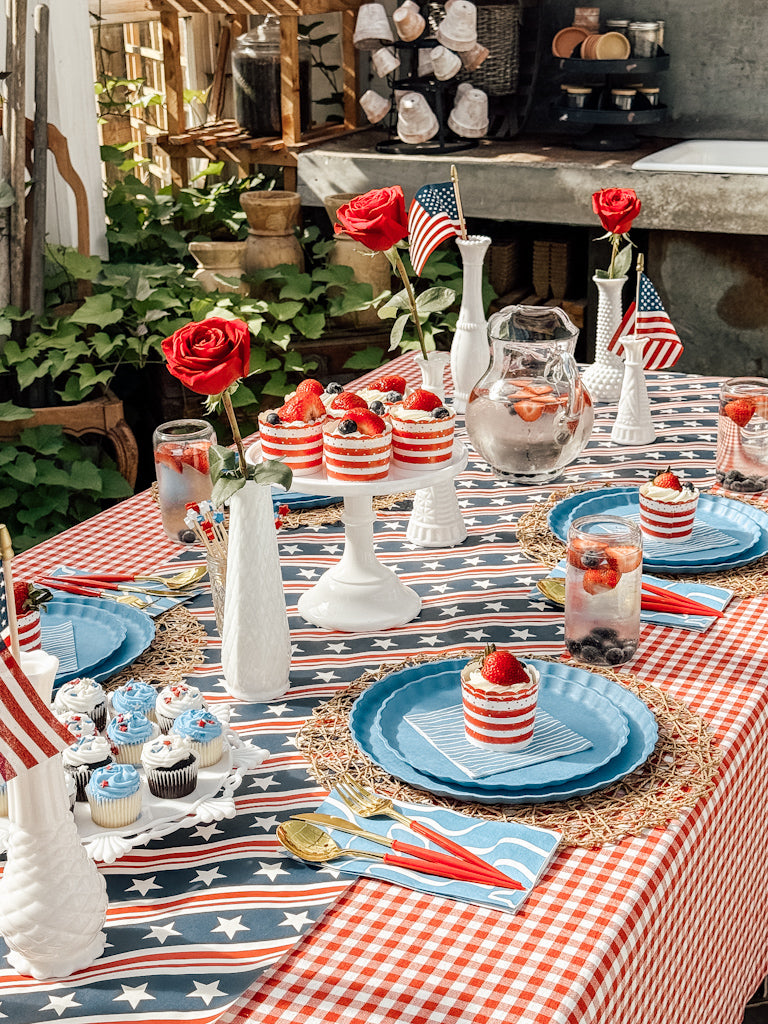 A festive celebration setup features Patriotic French Blue plates by Sophistiplate LLC, American flags, red roses, berry drinks, jarred desserts, cupcakes, and decorations on a red and white checkered tablecloth.