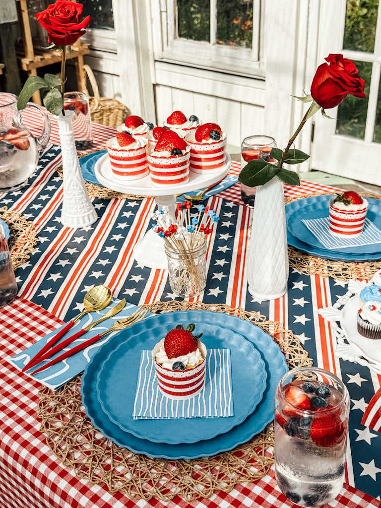 The table is set with Sophistiplate LLC's Patriotic French Blue plates, red, white, and blue desserts, cupcakes, strawberries, roses in vases, and berry drinks on star-spangled and checked tablecloths for a festive touch.