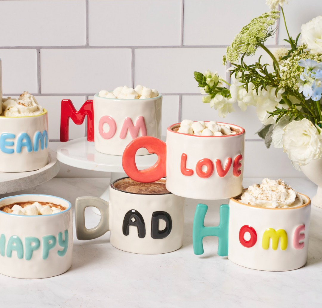 A Sophistiplate Ceramic Message Mug Mom, 14oz, filled with a hot drink and toppings, sits beside fresh flowers on a white counter.