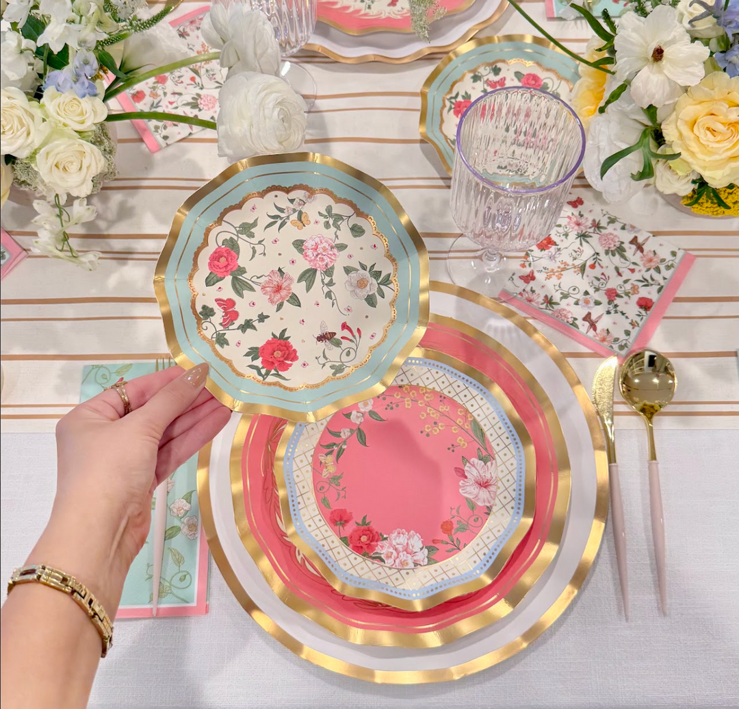 A hand holds a Rococo Table Setting designer disposable plate by Sophistiplate LLC, featuring a floral pattern over a pink and gold place setting with gold cutlery, a glass, and floral centerpieces on a white tablecloth.