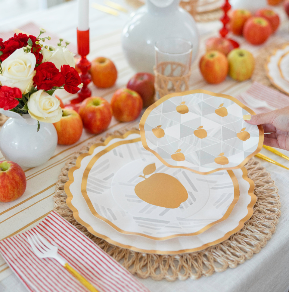 A beautifully decorated table showcasing apples, red and white flowers, striped napkins, and layered plates—one featuring a pear motif and another adorned with small apple patterns—displays the Rosh Tablescape for 16 Guests: Golden Apple by Sophistiplate LLC above the festive Rosh Hashanah setting. These heavy-duty paper plates bring both elegance and durability to your celebration.